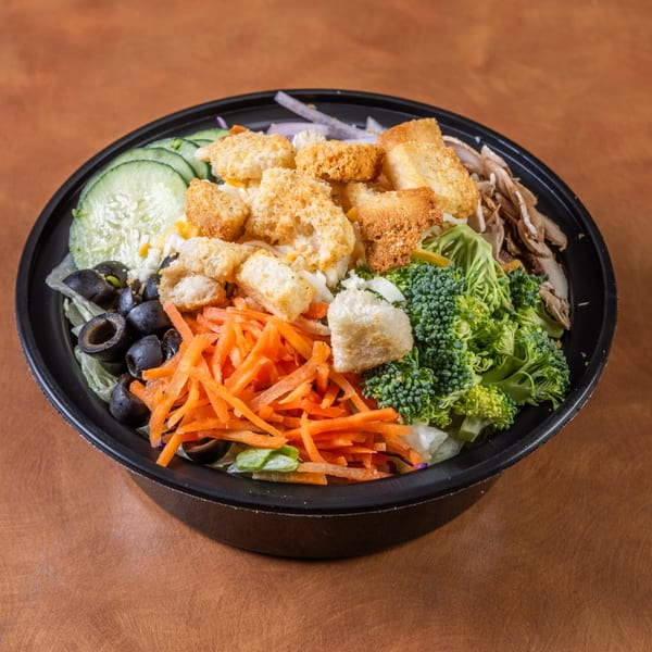 A black bowl filled with a fresh salad, including cucumbers, olives, carrots, broccoli, and croutons.