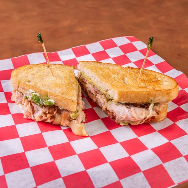 A grilled sandwich, cut in half, filled with meat, avocado, and cheese on red and white checkered paper.