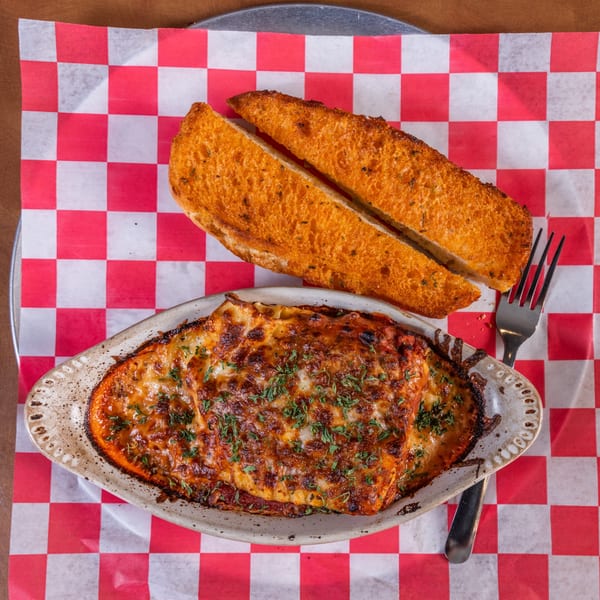 Baked lasagna with melted cheese and parsley, served with two slices of garlic bread and a fork.
